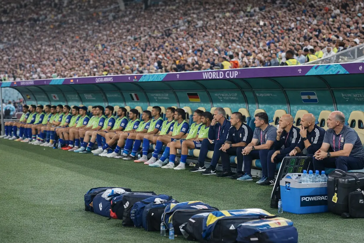 世界杯比赛现场替补席，替补球员与教练组并排就坐，场边摆放比赛装备